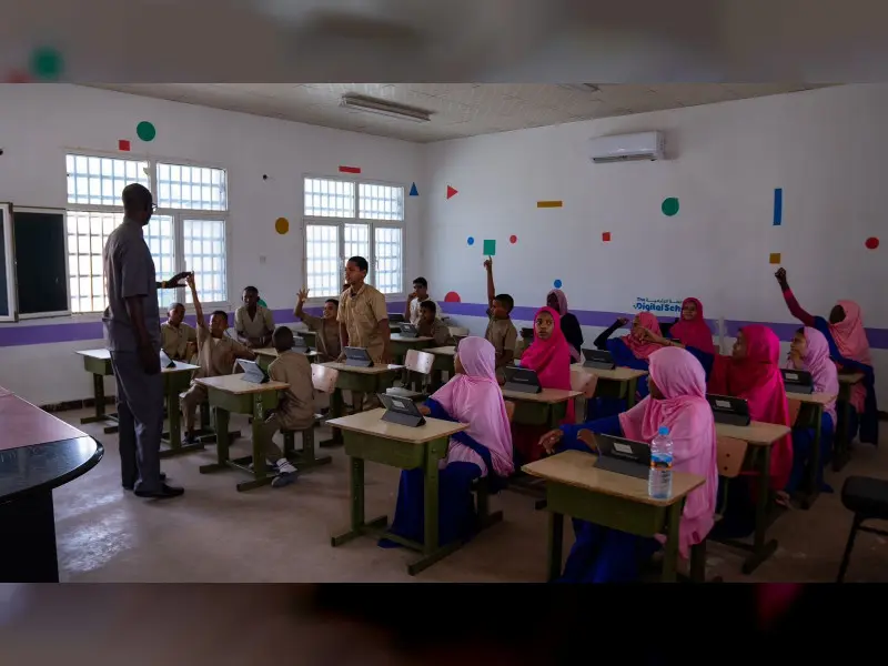 A classroom with students seated at desks, some in pink uniforms, eagerly raising hands. An ostaz stands at the front, engaging with the class. The room is adorned with geometric shapes, reflecting the innovative approach of Edtech in education across MENA.