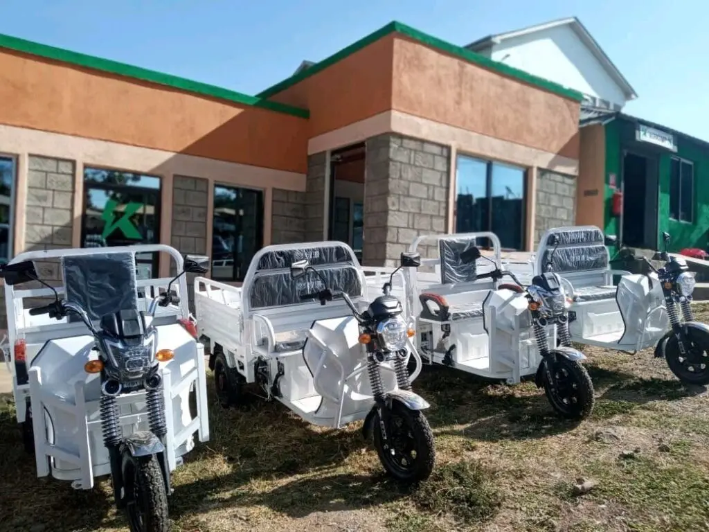 Four white motorized tricycles are parked outside a building with stone and orange walls.