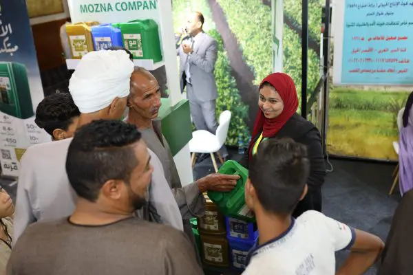 A group of people stands around a booth with colorful containers at an exhibit. A woman in a red headscarf is passionately discussing Mozna, an innovative Egyptian startup known for its organic fertilizer. Meanwhile, a man in a suit talks energetically in the background.