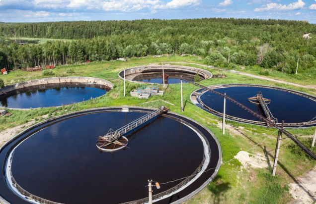 Aerial view of large circular wastewater treatment tanks surrounded by greenery and trees under a blue sky with scattered clouds.