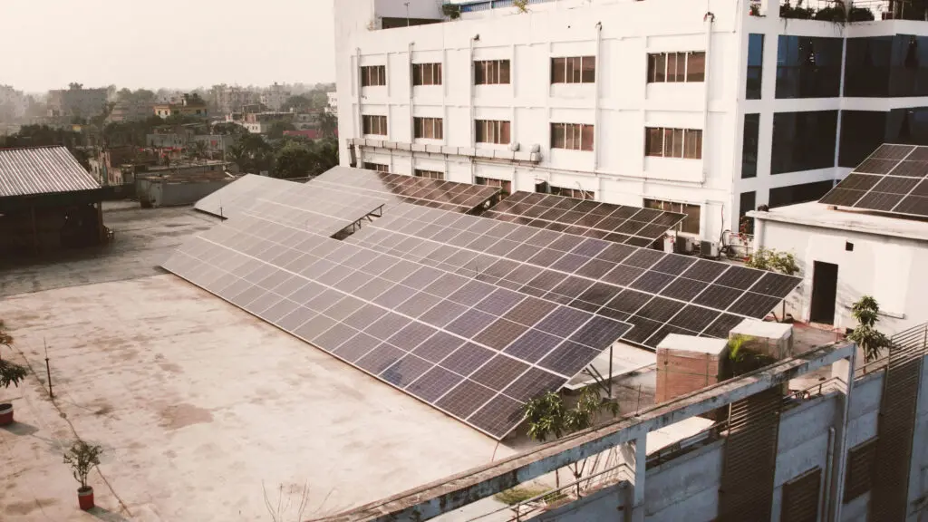 Solar panels glisten on the flat rooftop of a large white building, showcasing clean energy innovation in an urban area. Lush trees frame the scene, highlighting a sustainable future.