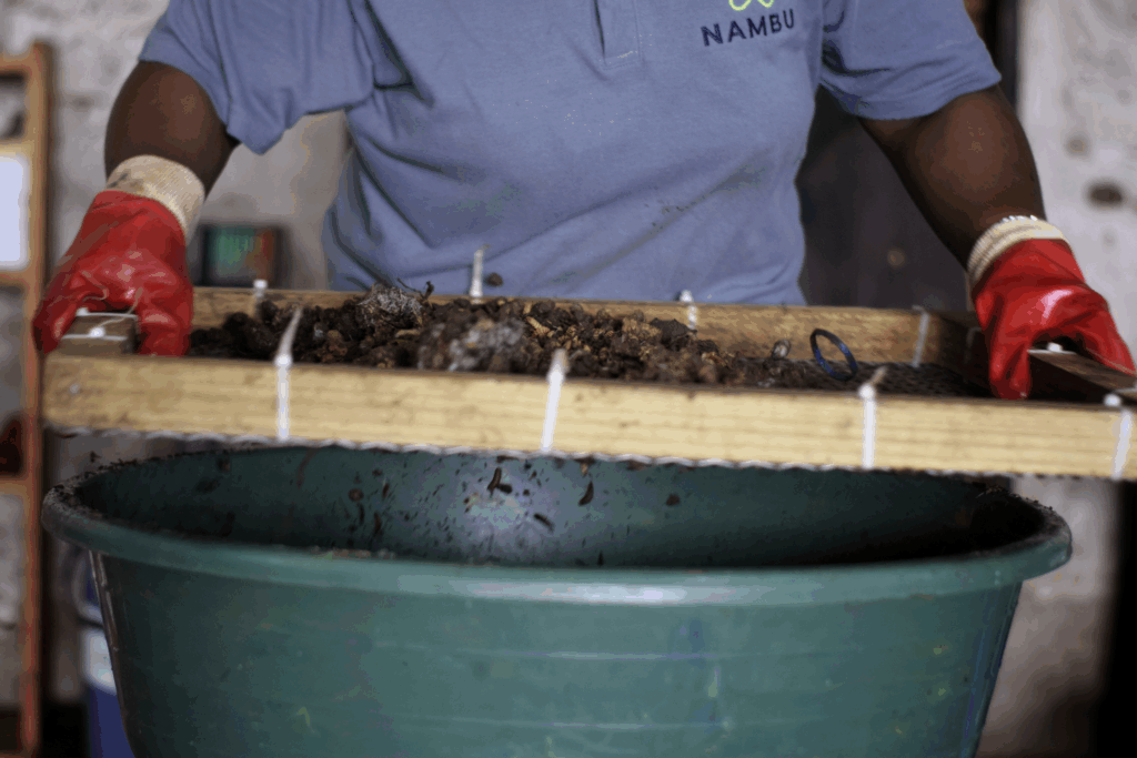 A person wearing red gloves sifts soil or organic material using a wooden frame over a large green container, showcasing nambu group's commitment to sustainability.