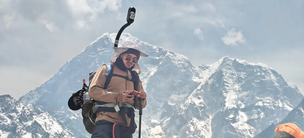 A person in hiking gear and sun hat uses a smartphone with a selfie stick, capturing memories against the backdrop of snow-covered mountains reminiscent of Everest, under a cloudy sky.