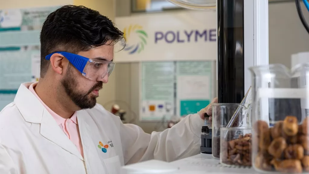 A man in a lab coat and safety goggles operates laboratory equipment with containers of samples nearby. The background features a sign that reads "POLYMERS," highlighting the lab's focus on Polymeron innovations and agro-solutions.